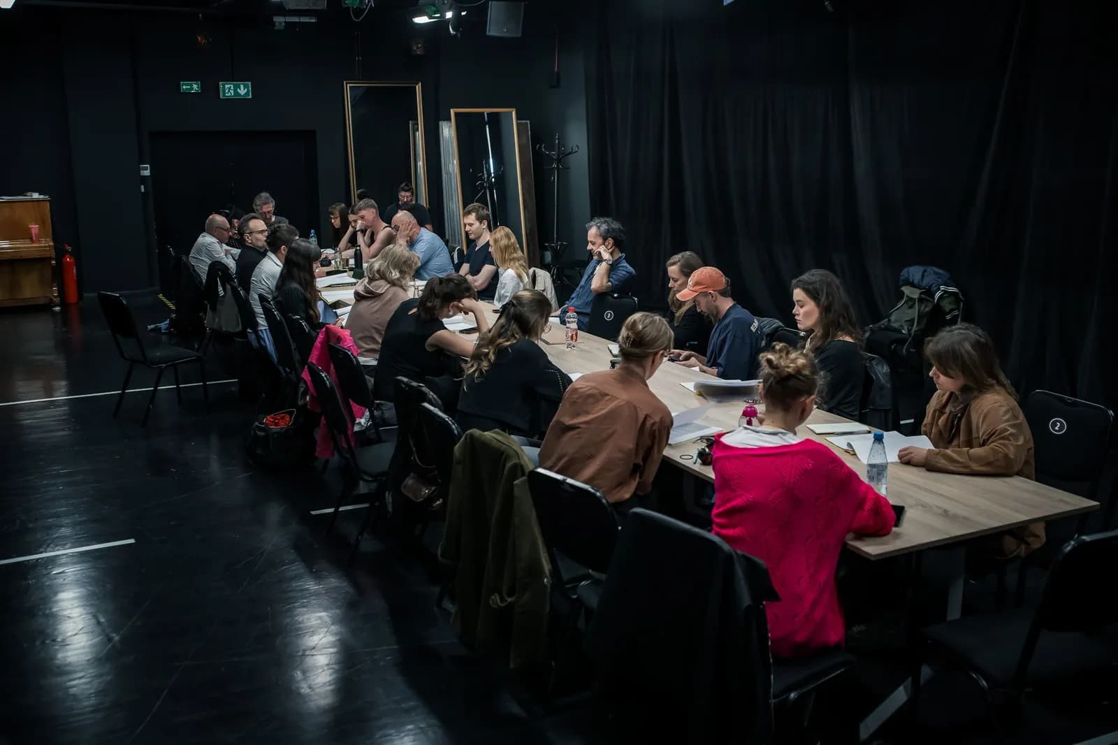 Grupa kilkudziesięciu osób siedzi przy długim stole w ciemnej sali teatralnej, czytając scenariusze i notatki podczas wspólnej próby.