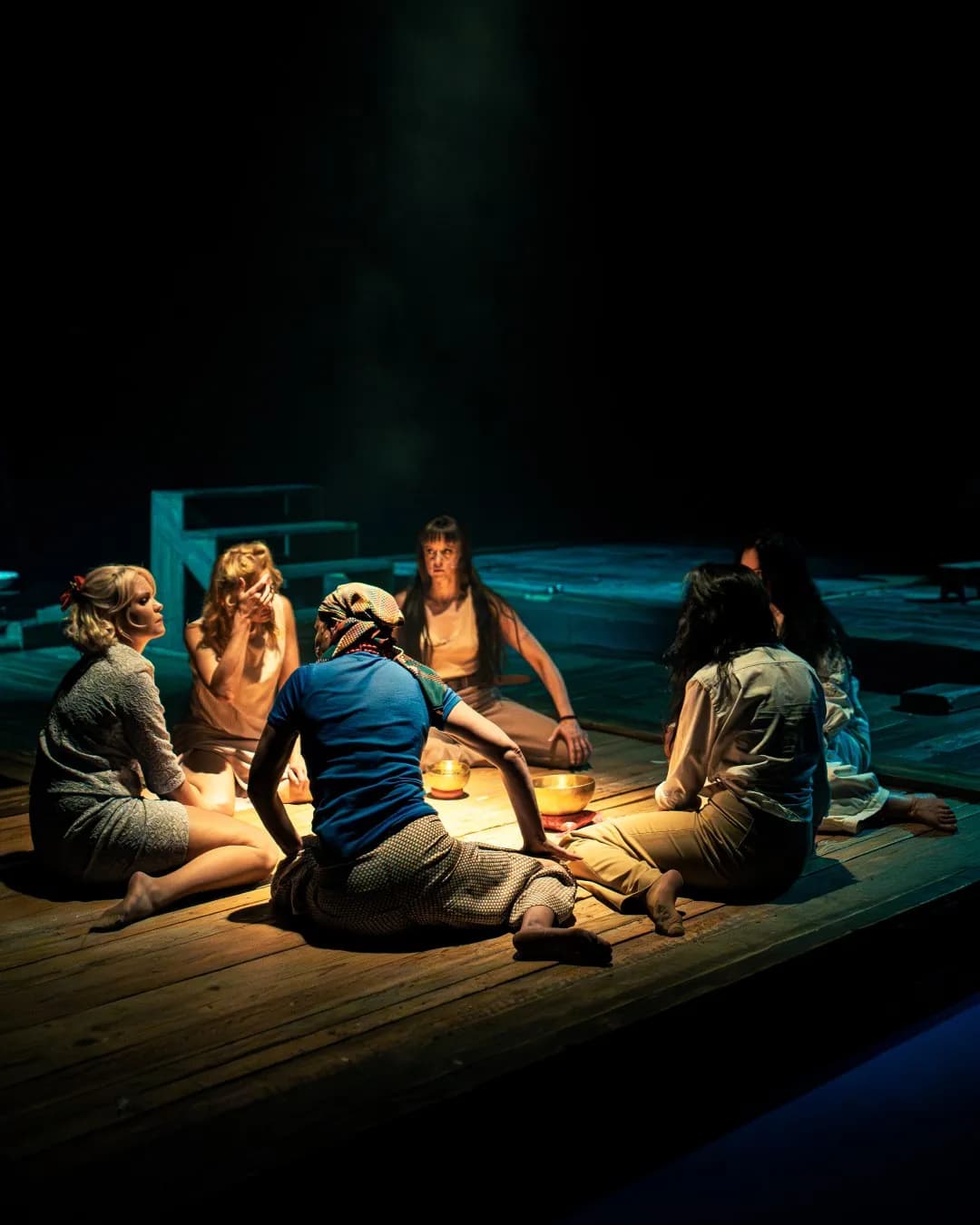 Sześć kobiet siedzi w kręgu na drewnianej scenie wokół metalowych mis. Kameralne spotkanie oświetla ciepłe, punktowe światło.