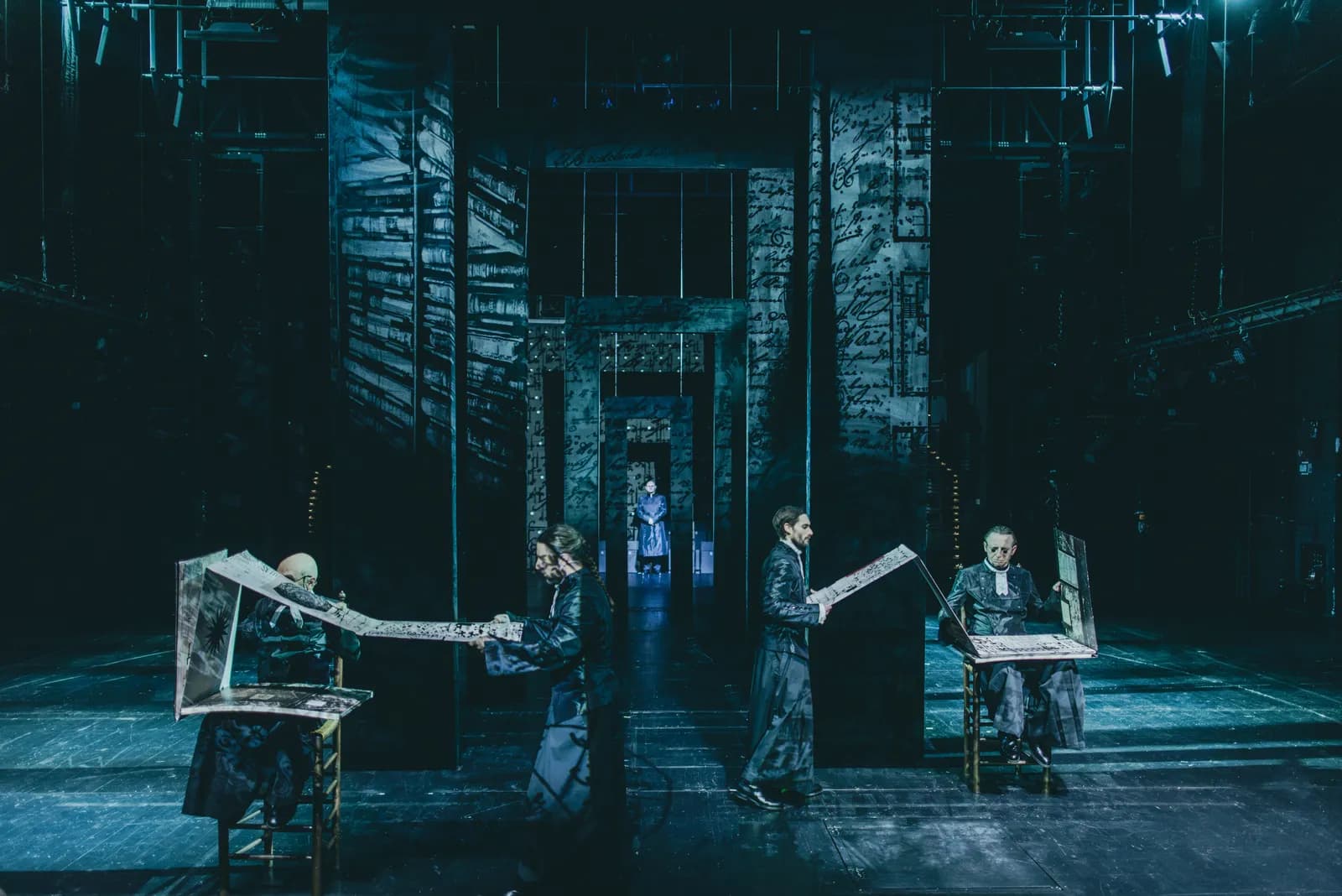 Na zdjęciu kilka osób na dużej scenie teatralnej, stojących przy pulpitach z długimi arkuszami papieru. W tle geometryczna scenografia z zapisami tekstowymi.