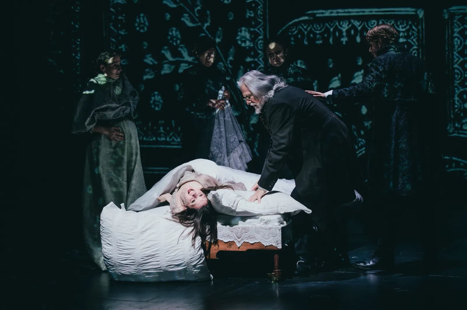Na zdjęciu kilka osób zgromadzonych wokół łóżka na scenie teatralnej. Jedna osoba leży na łóżku, inne stoją obok i pochylają się nad nią. Tło ciemne, projekcja dekoracyjna.
