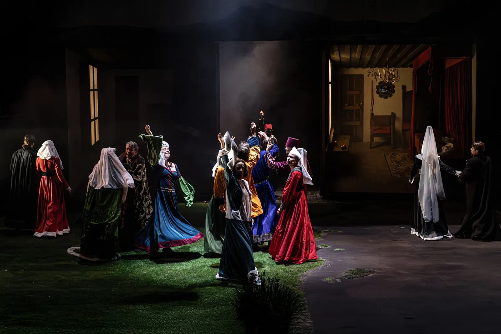 Grupa aktorów w barwnych, historycznych kostiumach tańczy na scenie teatralnej; w tle oświetlony pokój z łóżkiem i czerwoną kotarą.
