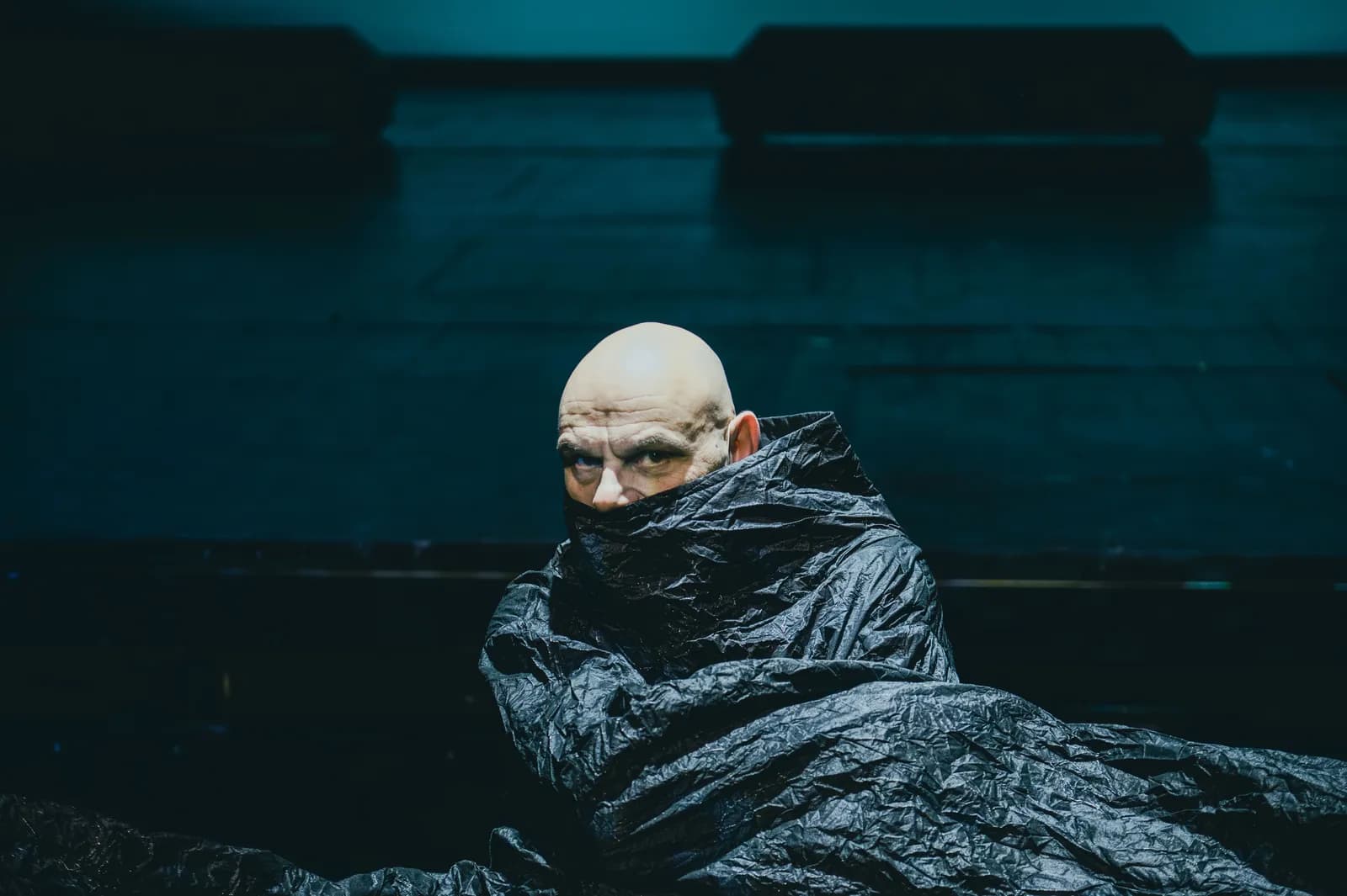 Na zdjęciu pojedyncza osoba (Robert Ninkiewicz) na scenie, owinięta ciemnym materiałem, z widoczną głową. Postać siedzi lub klęczy. Tło jednolite, niebiesko oświetlone.