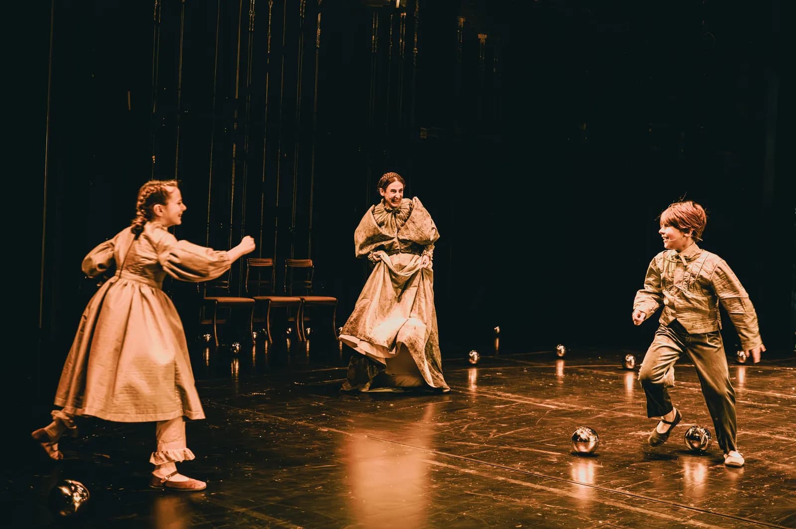 Na zdjęciu dwoje dzieci i dorosła osoba (Katarzyna Michalska) na scenie teatralnej. Dzieci stoją naprzeciw siebie, jak podczas zabawy. Dorosła osoba znajduje się pośrodku. Na podłodze leżą małe, kuliste obiekty.