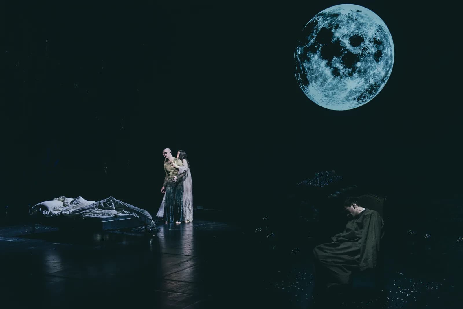 Na zdjęciu szeroki plan sceny teatralnej. Dwie osoby stoją obok łóżka po lewej stronie, jedna osoba siedzi po prawej. W tle widoczna projekcja księżyca na ciemnym niebie.