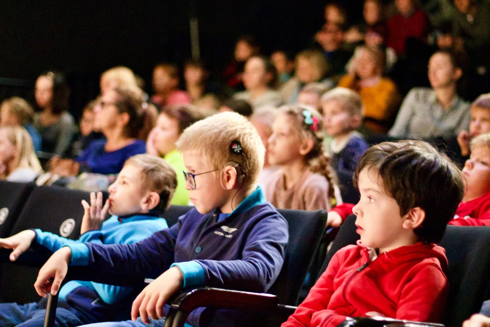 Dzieci siedzą na widowni podczas spektaklu. Patrzą uważnie przed siebie. Niektóre mają grube okulary lub aparat słuchowy.
