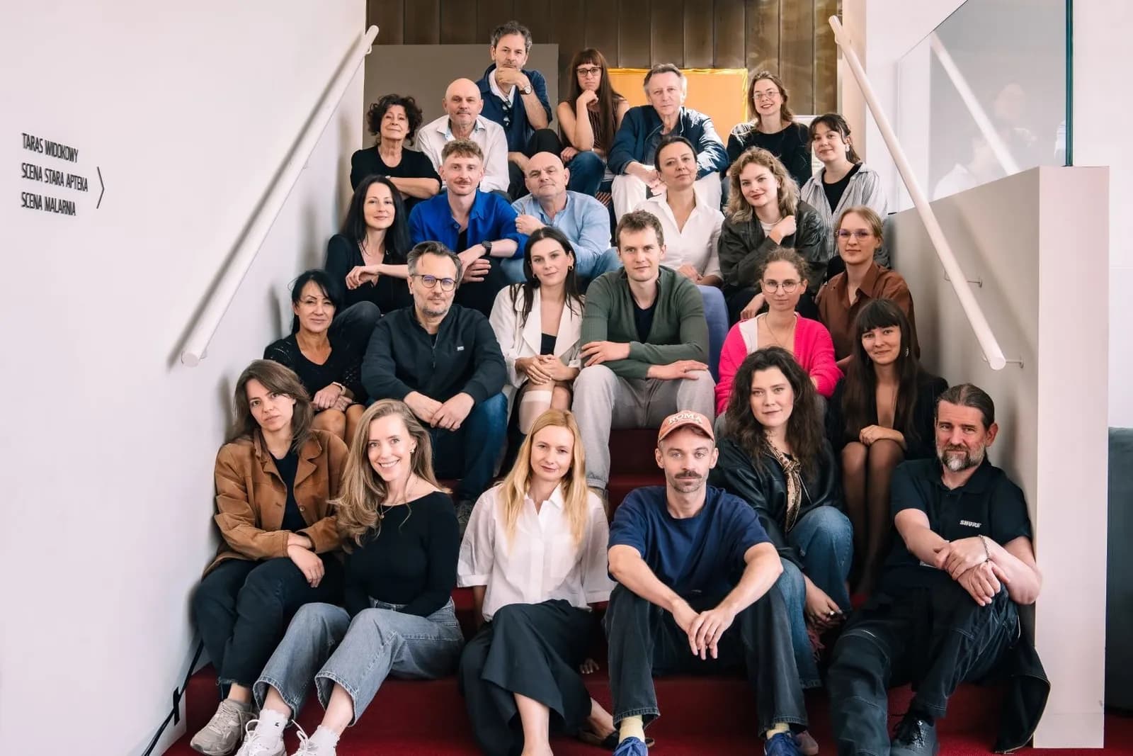 Grupa około dwudziestu pięciu osób pozuje do wspólnego zdjęcia na schodach we wnętrzu teatru. Większość siedzi, kilka osób stoi w tylnych rzędach.